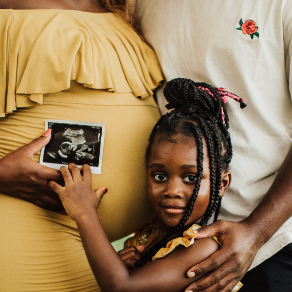 a pregnant woman, male partner, and young child stand with an ultrasound photo held up