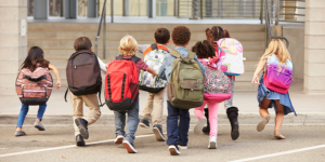 Children wearing backpacks run towards a school