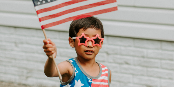 Young boy wearing star-shaped sunglasses and a red-white-and-blue outfit holds up an American Flag.
